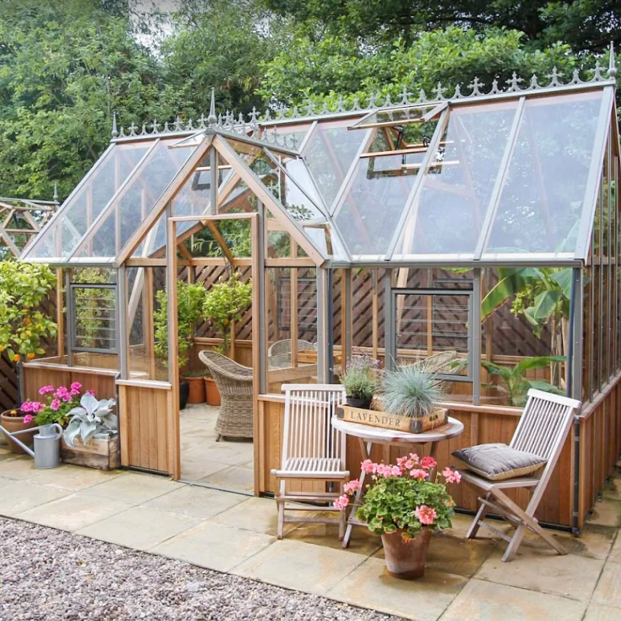 Alton Cedar Greenhouse with wooden frame and glass panels, surrounded by plants and a small table with chairs.