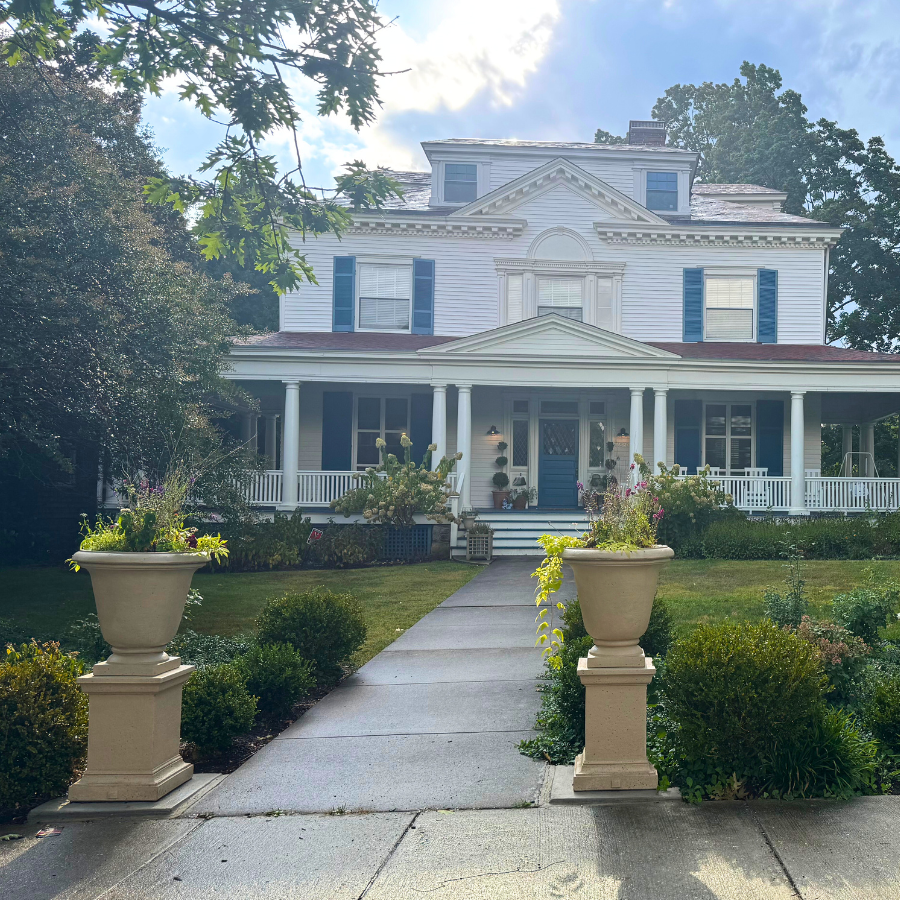 Large white house with blue shutters and a front porch, surrounded by greenery and planters in a cottage garden.