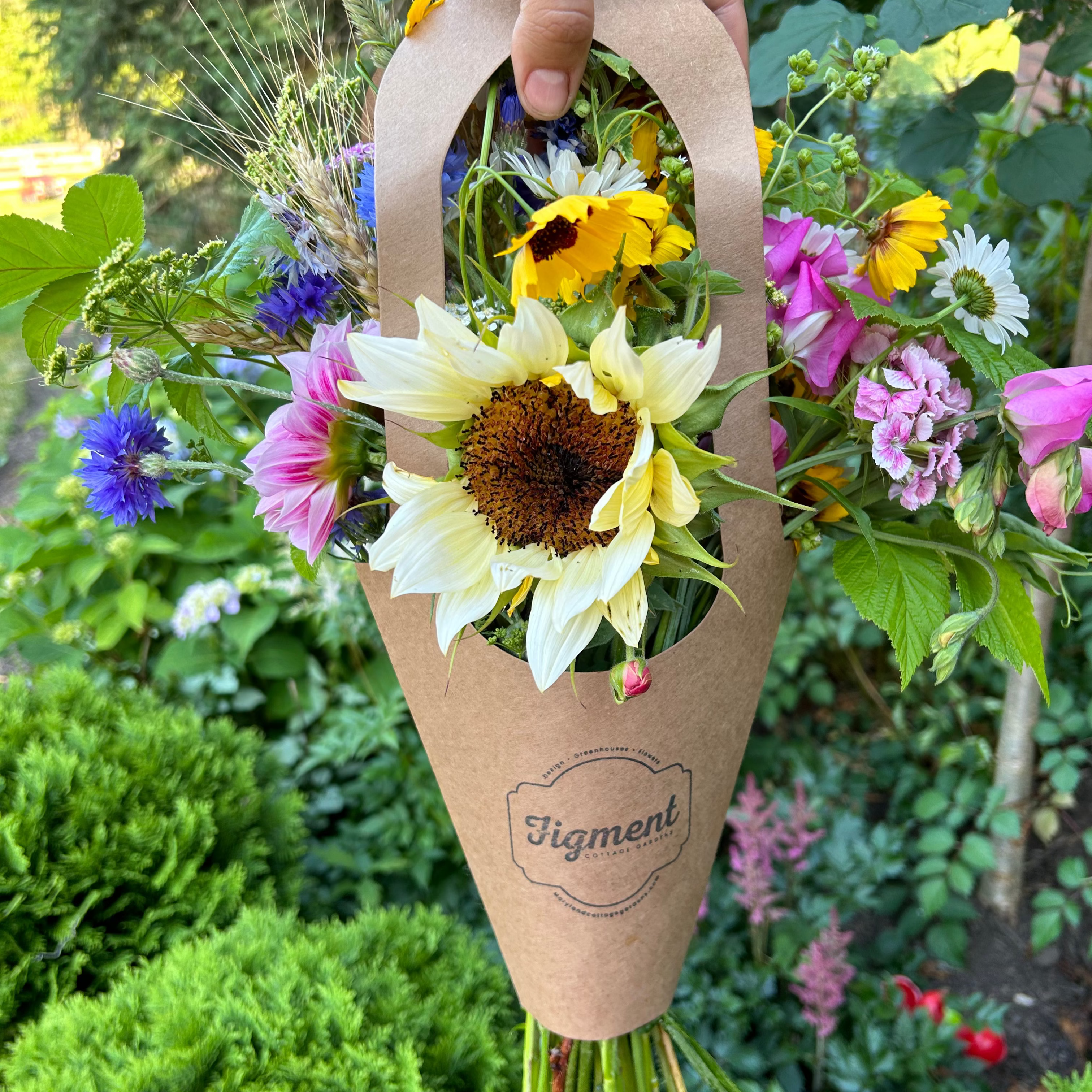 Bouquet of summer flowers and sunflowers in DIY bouquet flower tote with 'Figment' branding, held against a garden background.

