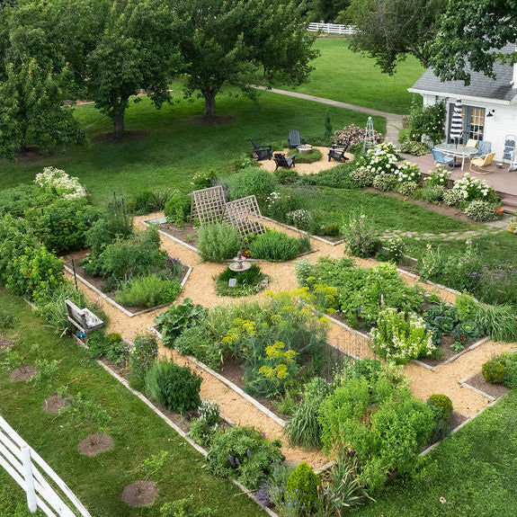 An aerial view of a large, professionally landscaped raised garden bed system with gravel paths and various green plants, situated next to a white house with a wooden deck.
